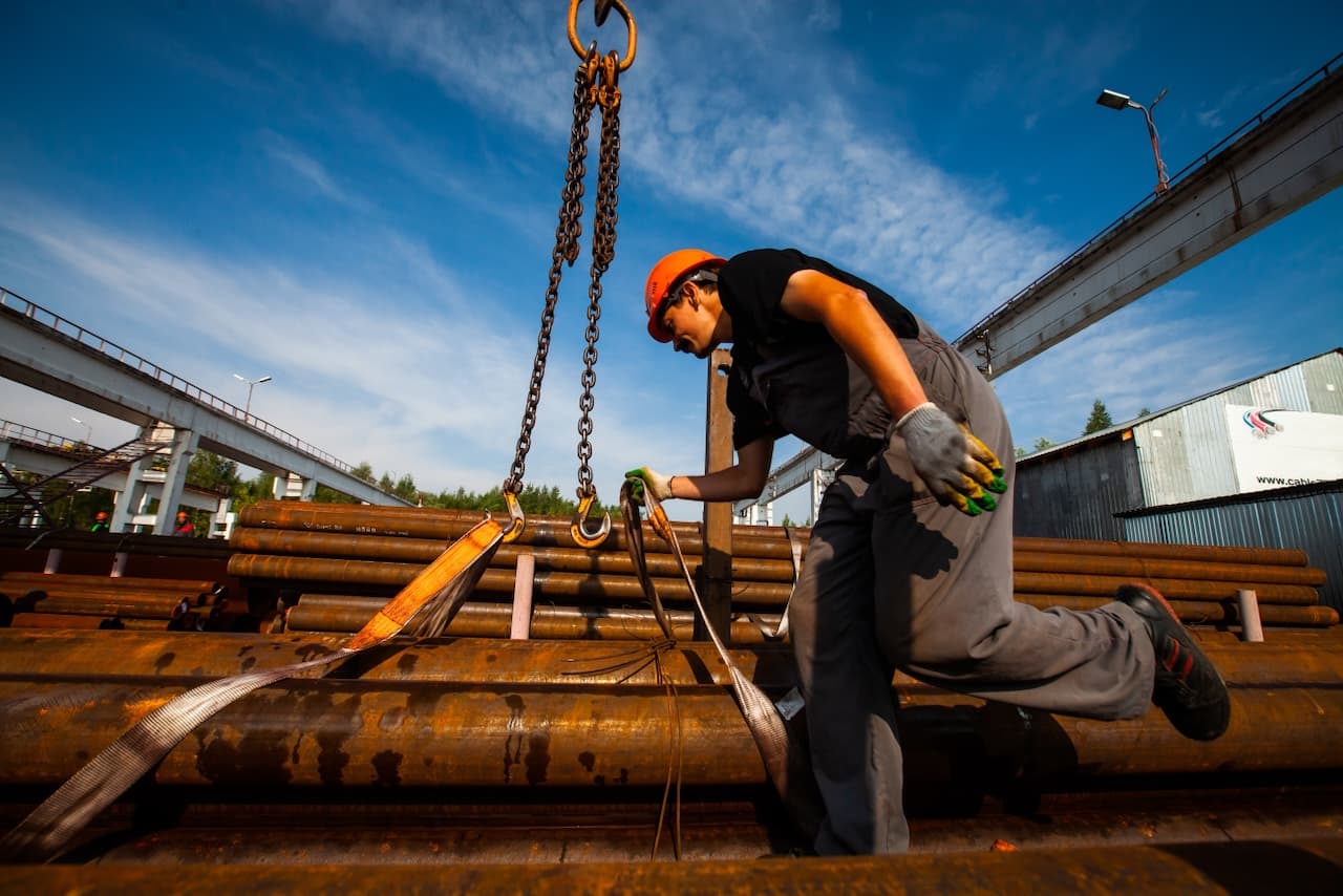 Trabajador ajustando las cadenas para levantar tubos de acero en un entorno industrial, destacando la dedicación y el cuidado en la manipulación de productos de alta calidad de MMTubería.